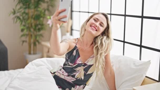 Νεαρή ξανθιά γυναίκα κάνει selfie από smartphone κάθεται στο κρεβάτι στο υπνοδωμάτιο - Πλάνα, βίντεο