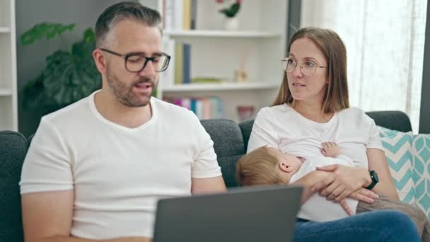 Az anya, apa és baba családja laptoppal otthon ül a kanapén - Felvétel, videó