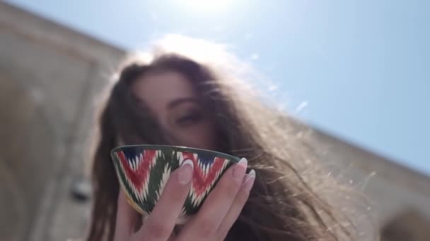 Una giovane donna con i capelli lunghi che tiene in mano una tazza di tè in ceramica dipinta con un ornamento uzbeko. Close Up, nozione lenta - Filmati, video