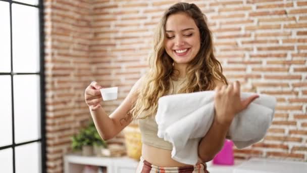Junge schöne hispanische Frau lächelt selbstbewusst und hält gefaltete Handtücher und Waschmittelbecher in der Waschküche - Filmmaterial, Video