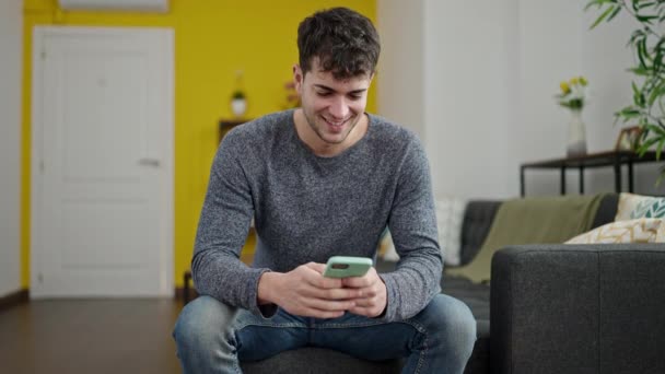 Junger hispanischer Mann sitzt mit Smartphone lächelnd zu Hause auf dem Sofa - Filmmaterial, Video