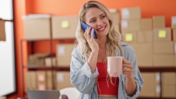 Nuori blondi nainen verkkokauppa liiketoiminnan työntekijä puhuu älypuhelimella juo kahvia toimistossa - Materiaali, video