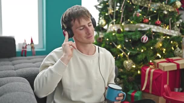 Młody biały mężczyzna słuchający muzyki pijący kawę świętujący święta w domu - Materiał filmowy, wideo