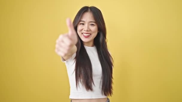 Fiatal kínai nő mosolygós magabiztos csinál rendben jel hüvelykujjával felfelé izolált sárga háttér - Felvétel, videó
