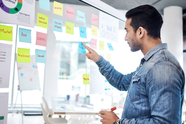 Creative man, sticky note and pointing on glass board for schedule tasks, planning or brainstorming at office. Male person or employee with plan, agenda or marketing strategy for startup at workplace. - Photo, Image