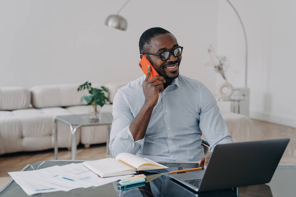 Happy African American businessman, making business call, enjoys conversation on mobile while working on laptop at office desk. - Фото, зображення
