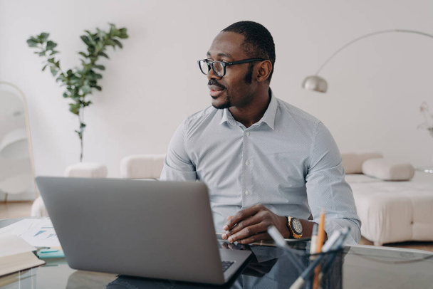 Pensive Afro-Amerikaanse zakenman, het dragen van een bril, werkt op laptop, nadenken project, zitten op kantoor. - Foto, afbeelding