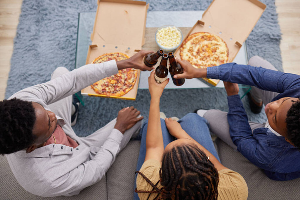 Cheers, beer and above of people with pizza, lunch celebration and relax together in a house. Happy, dinner and friends toasting with alcohol and food on the living room sofa to celebrate eating. - Foto, Imagen