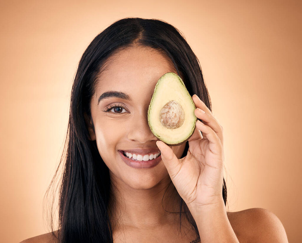 Happy, face and woman with avocado for skincare isolated on a brown background in studio. Portrait, fruit and model with food for nutrition, skin health and vegan diet, omega 3 or natural cosmetics. - Photo, image