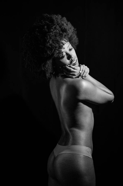 Sensuale donna afroamericana nuda con i capelli ricci che distoglie lo sguardo dalla spalla mentre si trova sullo sfondo nero. Fotografia in bianco e nero. - Foto, immagini