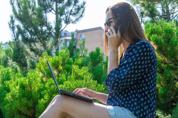 Kobieta pracuje na laptopie i rozmawia przez telefon komórkowy w parku miejskim. Kształcenie młodych studentów na świeżym powietrzu. Kobieta siedzi na zewnątrz i typy na komputerze. Miejsce pracy na przyrodzie w słoneczny dzień. Online. - Zdjęcie, obraz
