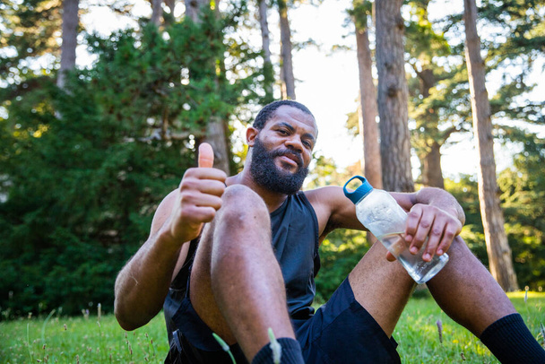 Uno sportivo seduto su un prato con una bottiglia d'acqua dà i pollici in su - Foto, immagini