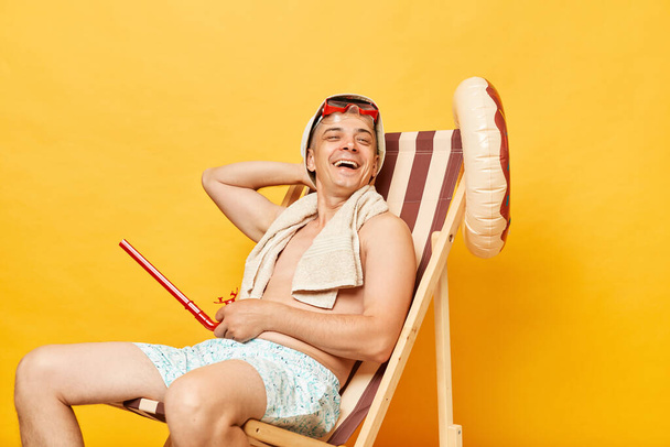 Relaxed satisfied smiling shirtless man sitting on deck chair isolated over yellow background enjoying his summer vacation laughing happily. - Valokuva, kuva