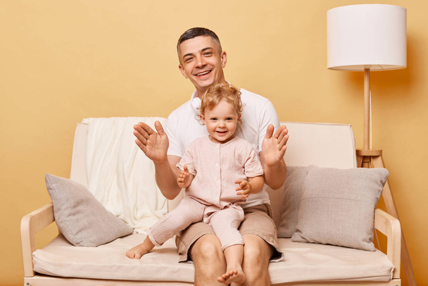 Attractive man with his daughter clapping hands and laughing while sitting on sofa tohgether playing games having fun at home expressing happiness. - Photo, Image