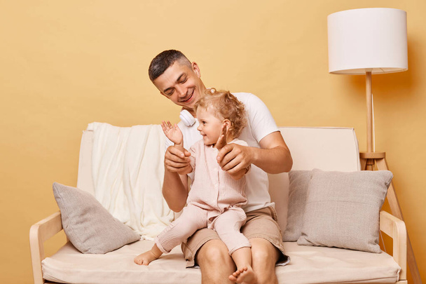 Father and baby girl smiling at camera while sitting on the sofa and clapping hands having fun together man spending weekend with his cute toddler daughter. - Φωτογραφία, εικόνα