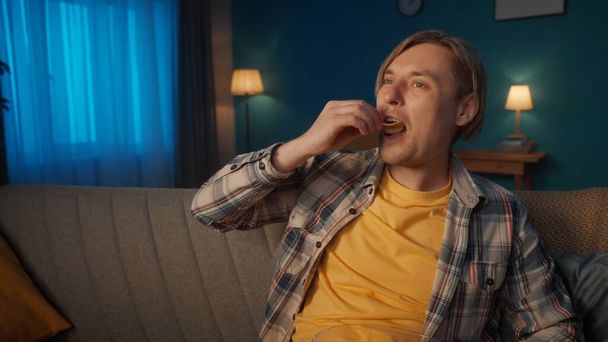 A man watches TV at home in the evening. A man sits on the couch and eats chips while watching a movie. Portrait of a man in the living room on the sofa close up - Foto, immagini