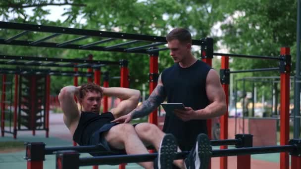 een fitnesstrainer met een tablet in zijn handen traint een jonge atleet die zijn buikspieren op straat pompt op een sportveld motivatie ondersteuning - Video