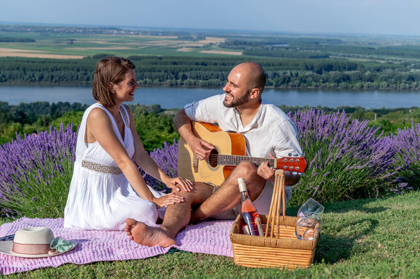 Zakochana młoda para lubi piknik na malowniczym lawendowym polu nad rzeką. Mąż gra na gitarze dla swojej uroczej żony.. - Zdjęcie, obraz