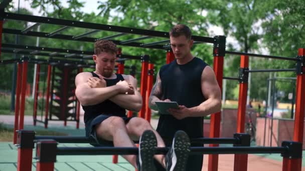 ένας γυμναστής με ένα tablet στα χέρια του εκπαιδεύει ένα νεαρό αθλητή αντλώντας τους κοιλιακούς του στο δρόμο σε αθλητικά γήπεδα υποστήριξη κινήτρων - Πλάνα, βίντεο
