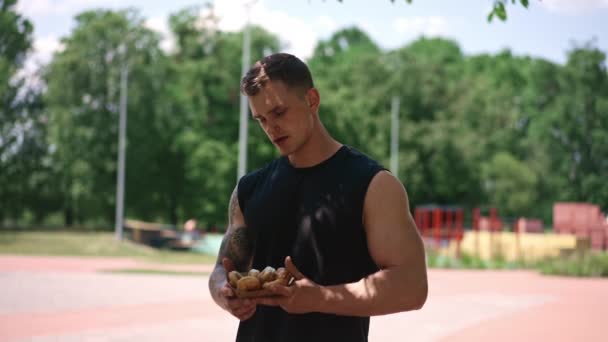 młody sportowiec po treningu trzymając słodkie pączki w rękach waha się, czy jeść im śmieciowe jedzenie - Materiał filmowy, wideo