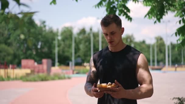 молодой спортсмен после тренировки держа в руках сладкие пончики колеблется, едят ли они нездоровую пищу - Кадры, видео
