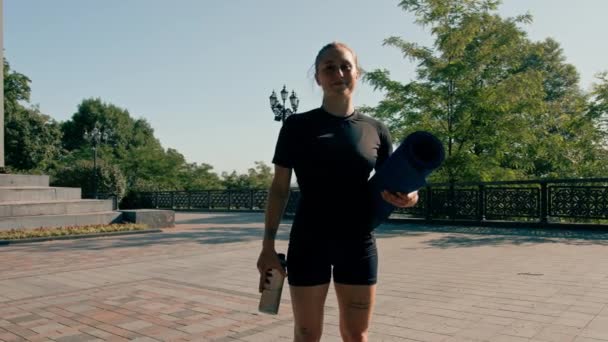 portrét mladé usměvavé sportovkyně držící v ruce kettlebell a láhev vody před zahájením cvičení venku v parku - Záběry, video