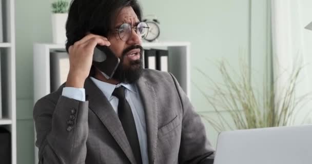 Een Indiase zakenman in een pak met een stropdas zit achter zijn bureau te bellen. Hij wordt erg boos terwijl hij aan de telefoon spreekt, legt de telefoon neer en houdt zijn hoofd vast van hoofdpijn.. - Video
