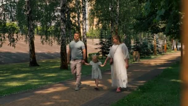 Klein schattig meisje in groene zomer jurk met ouders in het stadspark op wandeling zomer familie levensstijl kindertijd gelukkig gezin hand in hand - Video