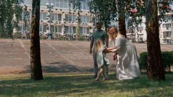Маленькая милая девочка в зеленом летнем платье с родителями в городском парке на прогулке летний семейный образ жизни Детство счастливая семья держась за руки - Кадры, видео
