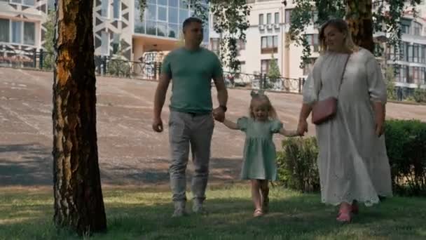 Niña linda en vestido de verano verde con los padres en el parque de la ciudad a pie estilo de vida familiar de verano Infancia familia feliz cogida de la mano - Imágenes, Vídeo