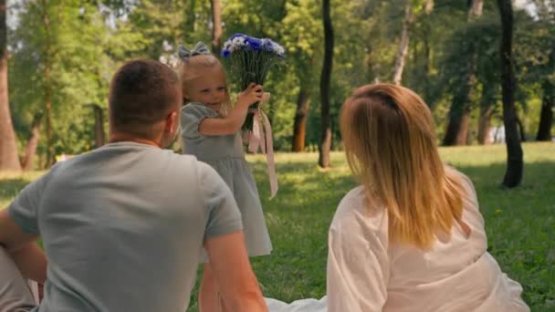una familia feliz descansa en la naturaleza en el parque una pequeña hija en un vestido da a su madre ramo de flores - Metraje, vídeo