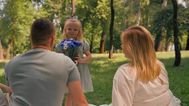 una familia feliz descansa en la naturaleza en el parque una pequeña hija en un vestido da a su madre ramo de flores - Imágenes, Vídeo