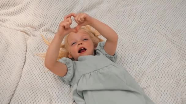 ritratto di una bambina felice sdraiata su una coperta nel parco Volto sognante di un bambino un bambino guarda il cielo mostrando il segno del cuore infanzia - Filmati, video