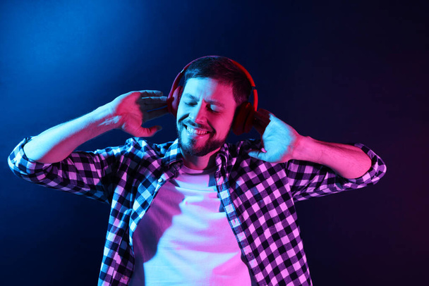 Щасливий чоловік в навушниках насолоджується музикою в неонових вогнях на темно-синьому фоні - Фото, зображення