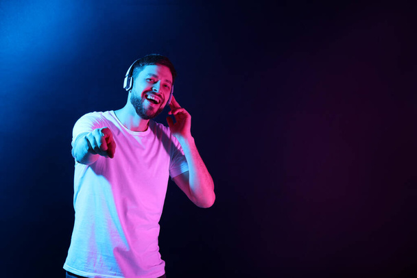Happy man in headphones enjoying music in neon lights against dark blue background. Space for text - Фото, изображение