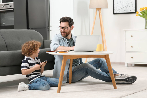 Man working remotely at home. Father using laptop at desk while his son playing with smartphone in living room - Foto, Bild