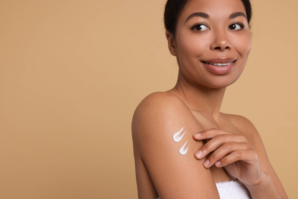Young woman applying body cream onto shoulder on beige background. Space for text - Фото, изображение
