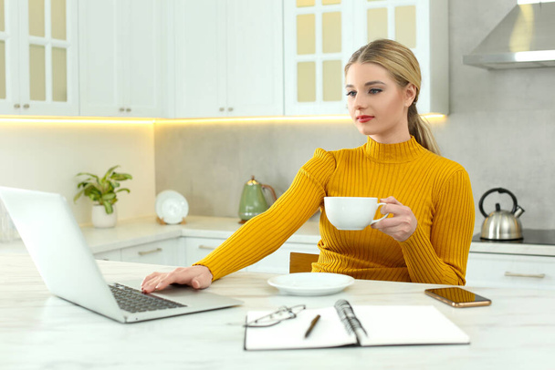 Home workplace. Woman with cup of hot drink looking at laptop at marble desk in kitchen - Φωτογραφία, εικόνα