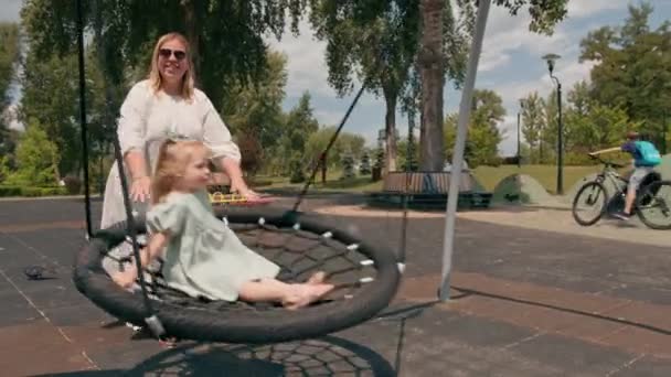 Klein schattig meisje in een groene zomer jurk met een strik op haar hoofd op de speelplaats moeder rijdt haar dochter op swing zomer jeugd - Video