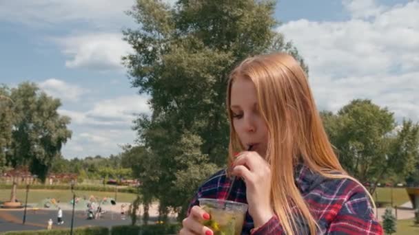 una joven camina por la ciudad en el verano, bebe un delicioso cóctel frío en muecas callejeras del sabor - Imágenes, Vídeo