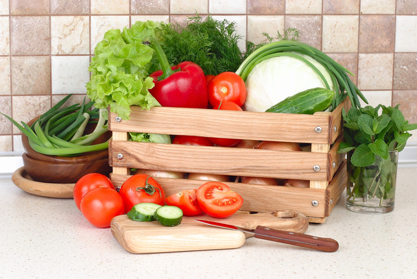 The fresh washed-up vegetables in a wooden box and the cut vegetables on a chopping board against modern kitchen. - Photo, Image