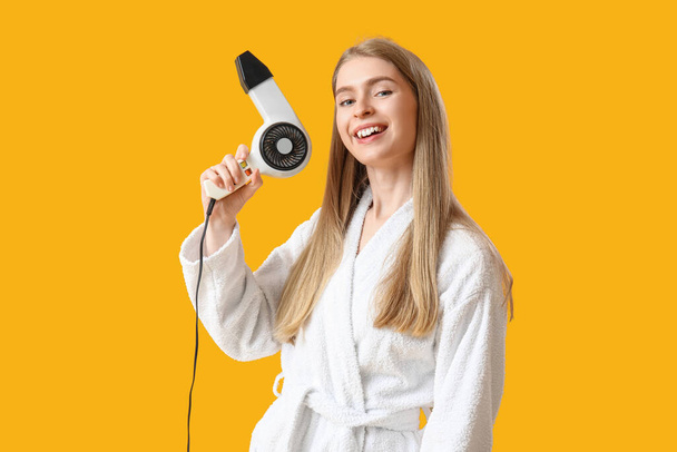 Young blonde woman with hair dryer on yellow background - Photo, Image