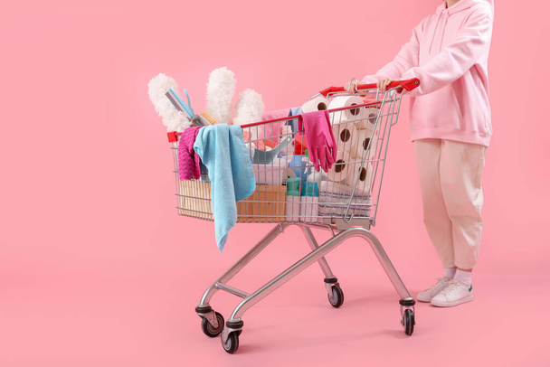 Vrouw met winkelwagentje vol schoonmaakspullen op roze achtergrond - Foto, afbeelding