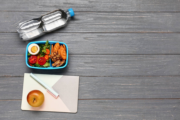 Bottiglia d'acqua, cancelleria e cestino per il pranzo con gustoso cibo su sfondo grigio in legno - Foto, immagini