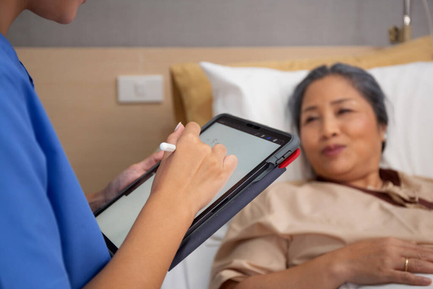 Closeup χέρια νοσοκόμα γραπτώς έκθεση έγγραφο σχετικά με συνταγογραφήσει ιατρική και υγειονομική περίθαλψη στο tablet, ενώ τον έλεγχο και τη διάγνωση με ηλικιωμένους ασθενείς στο νοσοκομείο, εξετάζοντας ασθένεια, ανώτερος άρρωστος. - Φωτογραφία, εικόνα