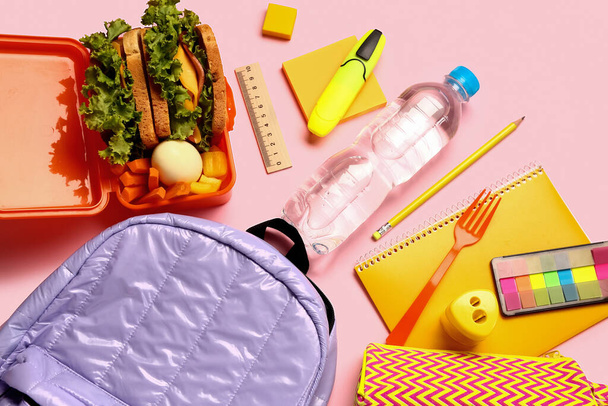 Hátizsák, írószer, üveg víz és uzsonnás doboz ízletes ételekkel rózsaszín alapon - Fotó, kép