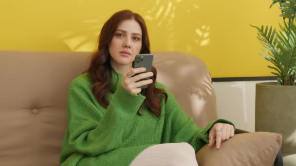 Красива молода жінка з темним красивим волоссям переглядає смартфон, сидячи на дивані вдома, кидаючи спокусливі погляди на камеру, посміхаючись. Високоякісні 4k кадри - Кадри, відео