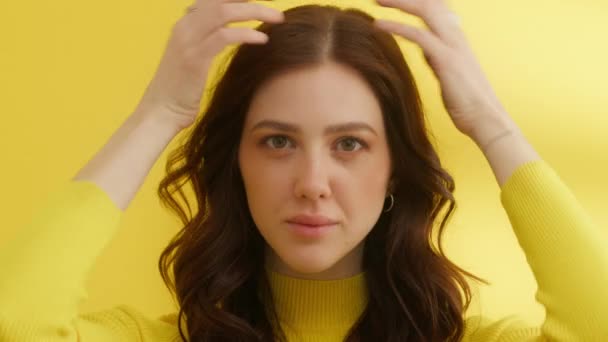 彼女のスタイルの髪に彼女の指に触れる美しい若いブルネットの中程度のショット。自信のある女性は黄色のセーターを着て、カメラを見てください。黄色の背景。高品質4k映像 - 映像、動画