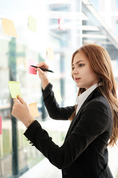 Beautiful female employee write down on colorful sticky notes manage list, concentrated biracial woman work on startup brainstorm collaborate plan on stickers on glass wall. High quality photo - Zdjęcie, obraz
