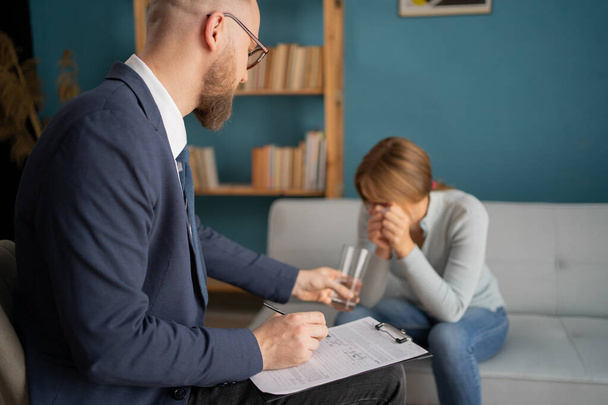 Depressed female patient sitting on psychotherapist couch as male doctor offers her a glass of water. Copy space - Photo, Image